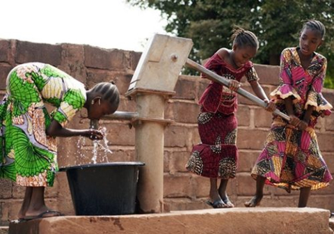 One young girl drinking from a public well while two other girls pump water